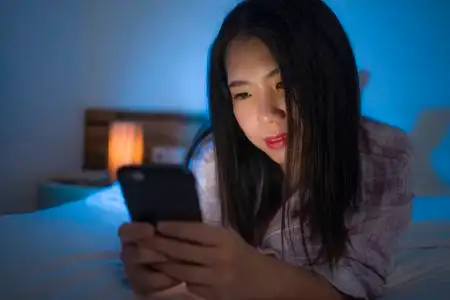 年轻美丽快乐甜美的亚洲韩国女人,穿着睡衣,晚上在床上享受手机应用