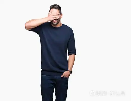 年轻的英俊男子穿着毛衣,在孤立的背景上微笑笑着,用手捂着眼睛,为