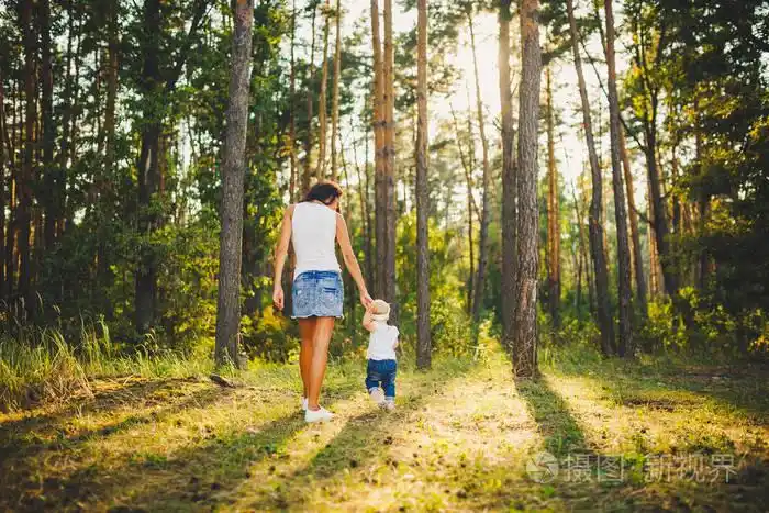 美丽而时髦的母亲在公园里牵着一岁的女孩的手, 在高大的针叶树的背景